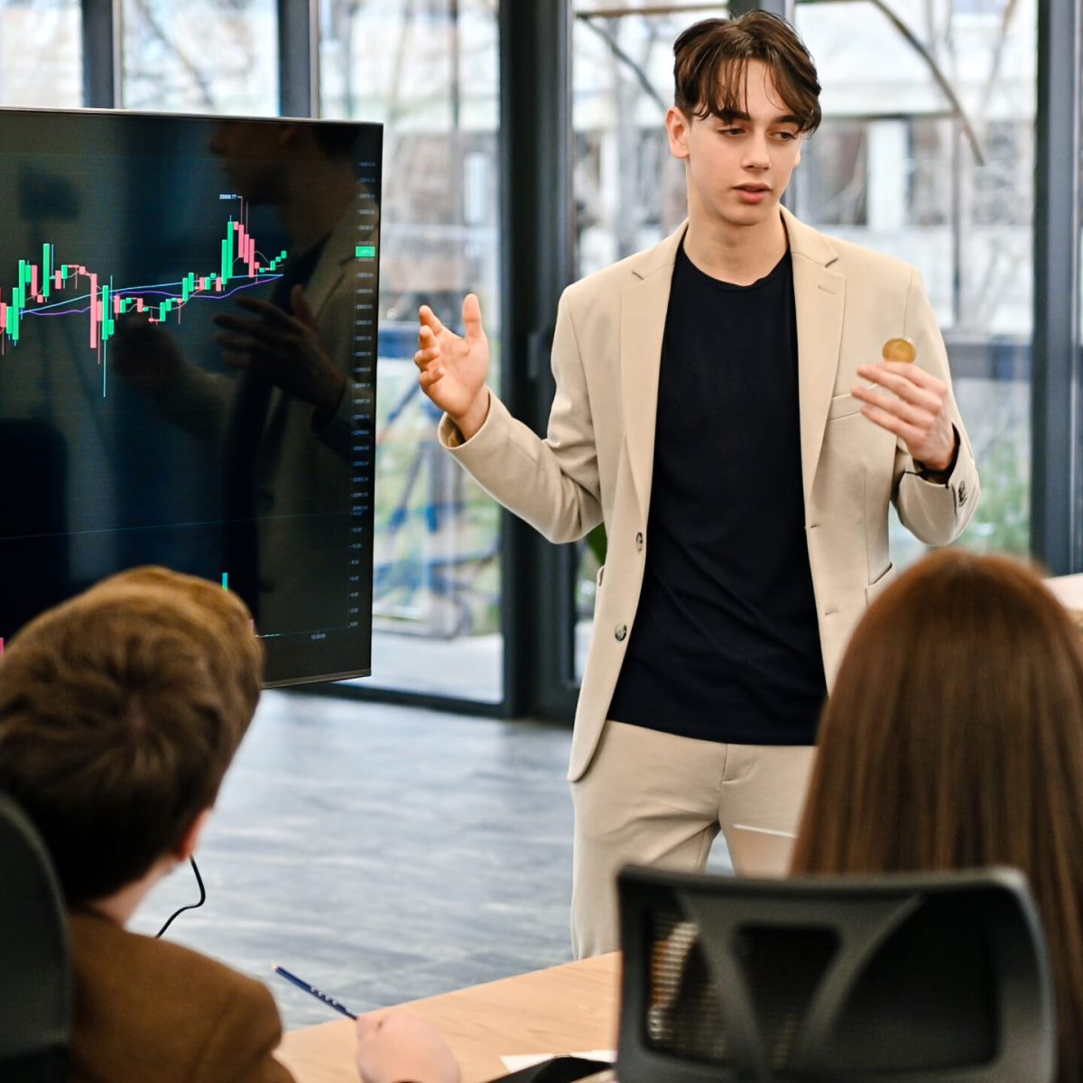 Young worker leading business meeting in an office, discussing the topic of cryptocurrencies with other workers using a big display with currency rate and physical coin